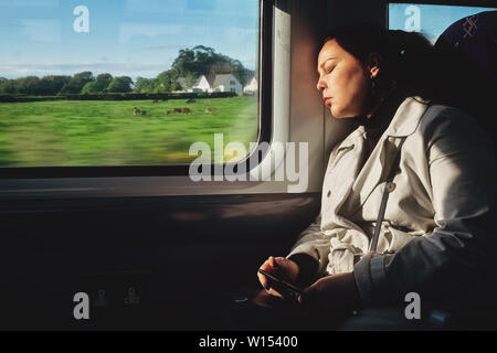 A young woman is sleeping in a train carriage holding a telephone. Outside the carriage window is a rural landscape Stock Photo