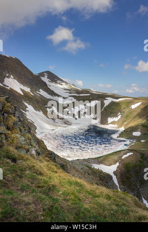 Beautiful, framed, sunlit view from Lakes peak on Rila mountain on The Eye lake, one of famous seven Rila lakes, still covered by broken ice in June Stock Photo