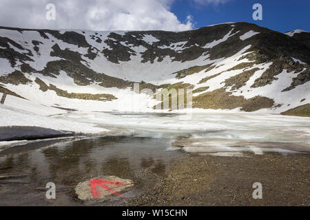 Low angle view of the tear lake partially frozen and covered by snow, impressive Rila mountain range and red arrow on a rock as a trail mark Stock Photo
