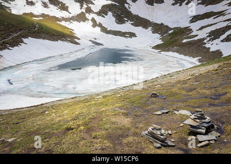 The Tear lake, one of famous seven Rila lakes, covered by colorful broken ice and snow, foreground piles of rocks and sunlit highlands Stock Photo