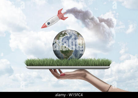 Woman's hand holding digital tablet with grass and planet Earth on screen, and rocket in air above leaving smoke trail behind. Stock Photo