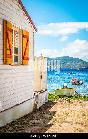 Wooden side wall of a white house with yellow and red window shutter. In the background a bay of water with a motor boat on the beach. Terre-de-Haut, Stock Photo