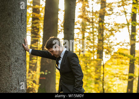 Young businessman in a suit leaning with his arm on a tree with his head bend down in meditation or disappointment. Stock Photo