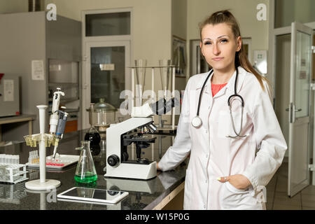 Young, Caucasian white, female laboratory technician in white robe with stethoscope standing next to compound microscope, laboratory, equipment and gl Stock Photo