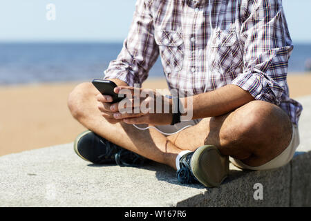 Mid section portrait of contemporary young man using smartphone sitting on concrete block by sea, copy space