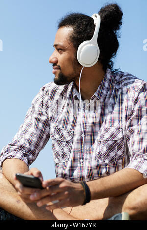 Side view portrait of contemporary Latin-American man chilling on beach listening to music while sitting cross legged, copy space Stock Photo