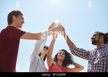 Low angle portrait of friends clinking beer bottles against blue sky in Summer, copy space Stock Photo