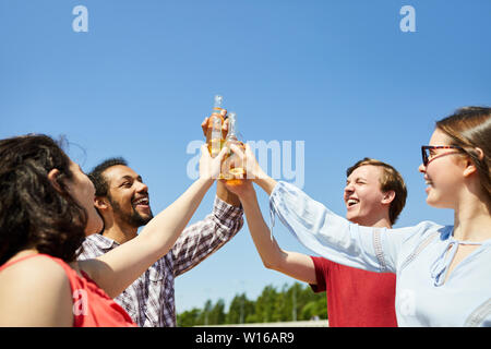Waist up portrait of happy young friends clinking beer bottles against blue sky in Summer, copy space Stock Photo
