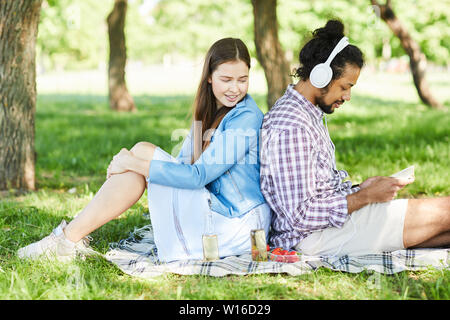 Side view portrait of young interracial couple sitting back to back outdoors during Summer picnic, copy space Stock Photo