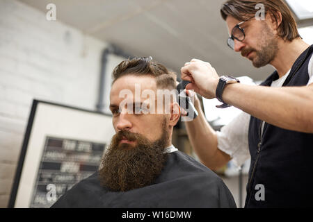 Brutal guy in modern Barber Shop. Hairdresser makes hairstyle a man with a long beard. Master hairdresser does hairstyle with hair clipper Stock Photo