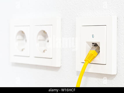 Yellow network cable in wall outlet for office or private home lan ethernet connection with power outlets flat view on white plaster wall background - Stock Photo