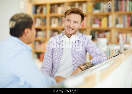 Content pensive tattooed man with headphones on neck sitting at table and discussing startup ideas with colleague in library Stock Photo