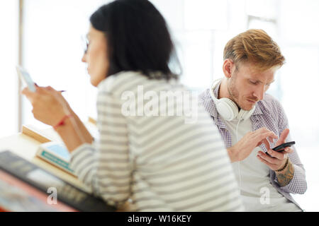 Serious modern students addicted to gadgets sitting at desk with books and scrolling in smartphones Stock Photo