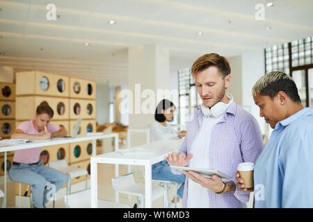Pensive young multi-ethnic bookstore colleagues in casual shirts standing against high tables for customers and using tablet while discussing sales of Stock Photo