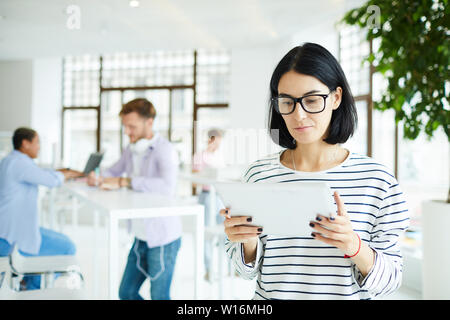 Serious pensive young female freelancer in eyeglasses standing in modern room and analyzing online sources on tablet Stock Photo