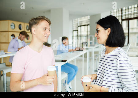 Positive confident young students in casual wear standing in campus hall and drinking coffee while discussing joint project Stock Photo