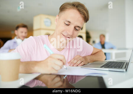 Positive confident young guy sitting at desk in library and making notes in workbook while preparing scientific research project Stock Photo