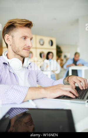 Serious focused handsome young man with headphones on neck sitting at table and using laptop while searching for online information Stock Photo