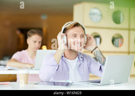 Positive excited handsome young guy with tattoo sitting at desk in library and adjusting headphones while preparing to work on project Stock Photo
