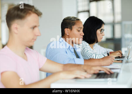 Group of concentrated young multi-ethnic people sitting at table in row and working on projects in co-working space Stock Photo