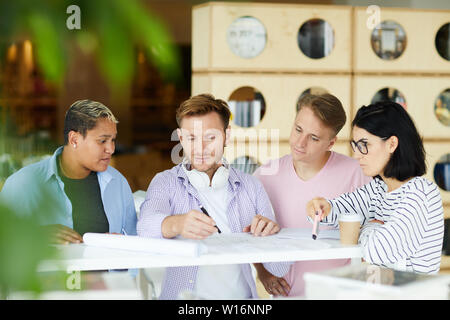 Group of young multi-ethnic architects standing at high table and working with blueprint while discussing design together Stock Photo