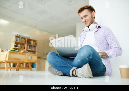 Cheerful positive hipster student guy with headphones on neck sitting with crossed legs on floor and using laptop while surfing net Stock Photo