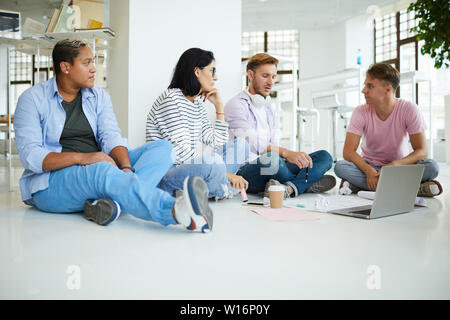 Group of thoughtful young interracial colleagues in casual outfits sitting on floor in library and working on creative project together Stock Photo