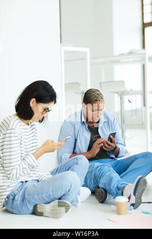 Concentrated mobile young multi-ethnic students sitting on floor and using smartphones while scrolling through social media Stock Photo