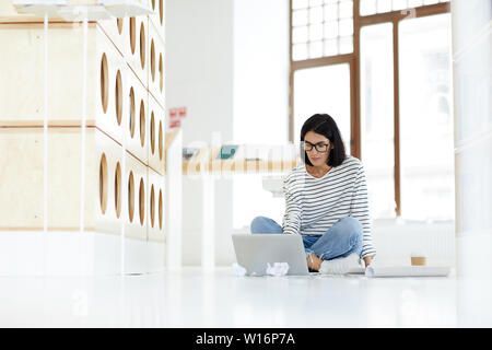 Concentrated modern female marketing specialist in eyeglasses sitting with crossed legs on floor in library and using laptop while checking data in in Stock Photo