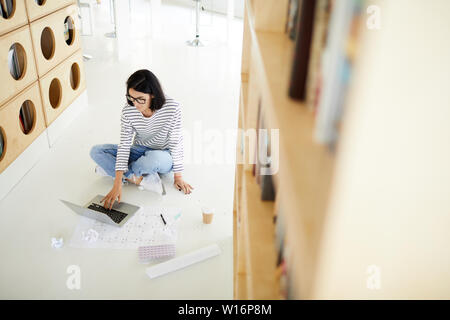 Above view of concentrated female flat designer sitting on floor with blueprint and crumpled papers and working on sketch in library Stock Photo
