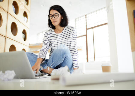 Serious intelligent young lady with black hair sitting with crossed legs on floor and using laptop while planning work schedule Stock Photo