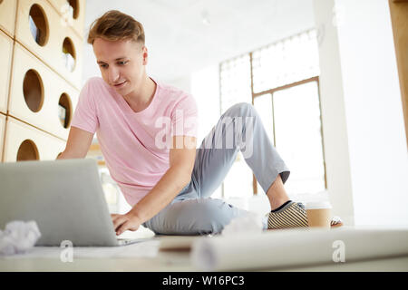 Content handsome student guy in casual clothing sitting on floor in library and using laptop while browsing internet Stock Photo