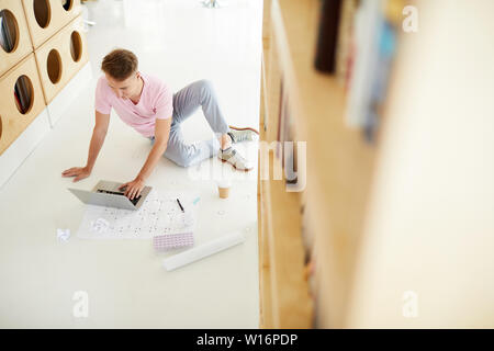 Content curious young male architecture student in casual clothing sitting on floor with blueprint and crumpled paper pieces in library and using lapt Stock Photo