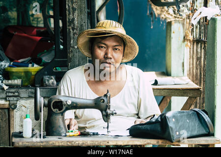 Yangon, Myanmar - March 2019: Asian man in straw hat behind retro mechanical sewing machine in the street tailor shop. Stock Photo