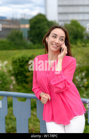 Happy relaxed attractive young woman using a mobile to chat to her friend as she leans on an metal railing smiling and looking to the side Stock Photo