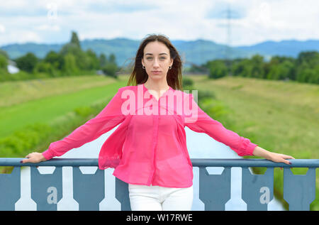 Stylish young woman in colorful pink blouse posing above a river leaning on the bridge railings looking solemnly at the camera Stock Photo