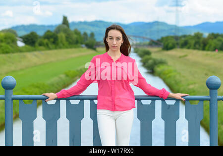 Stylish young woman in colorful pink blouse posing above a river leaning on the bridge railings looking solemnly at the camera Stock Photo