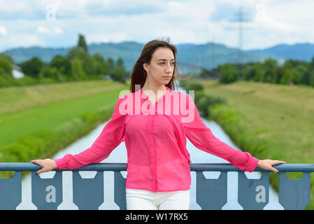Stylish young woman standing on a bridge thinking resting her arms on the railings and looking down at the ground with a pensive serious expression ag Stock Photo