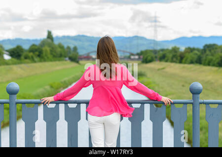 Young woman standing looking out over a rural river in lush green countryside leaning on the blue metal railings of the bridge Stock Photo