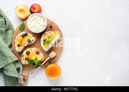 Ricotta and fruit bruschetta. Toasted baguette bread with ricotta cheese, peach, honey and blueberry on wooden board, concrete background. Top view, c Stock Photo