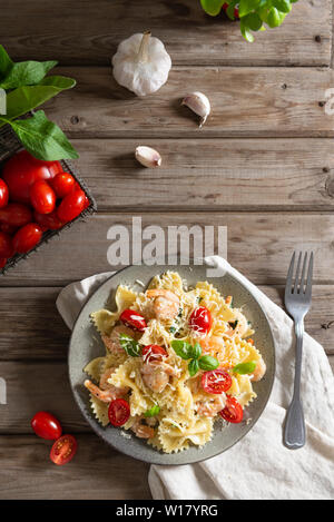 Italian pasta farfalle in a creamy garlic sauce with shrimps, cherry tomatoes and basil on a plate, top view. Copy space for text. Rustic Stock Photo
