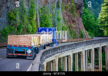 Big rig blue long haul semi truck with two flat bed semi trailers transporting industrial lumber boards running on the winding road with bridge around Stock Photo