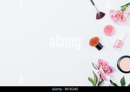 Pastel flat lay composition of makeup products and accessories next to delicate pink flowers peonies on white background with copy space.