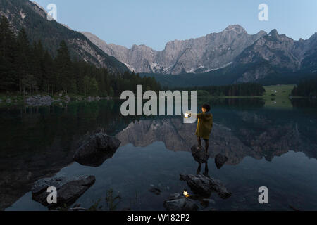 Boy in yellow rain jacket standing on the rock and holding a luminous hand lantern at scenic summer lake landscape in twilight, Laghi di Fusine, Italy Stock Photo