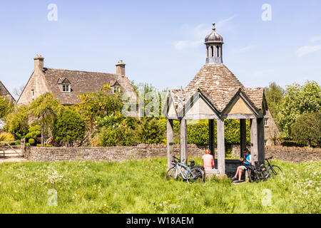 Maytime in the Cotswolds - Two female cyclists in the 1874 pumphouse by old stone cottages facing the village green in the village of Farmington UK Stock Photo
