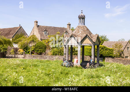 Maytime in the Cotswolds - Two female cyclists in the 1874 pumphouse by old stone cottages facing the village green in the village of Farmington UK Stock Photo