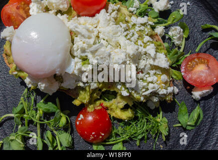 Smashed Avocado on sourdough toast topped with feta cheese and poached egg with salad on black plate Stock Photo