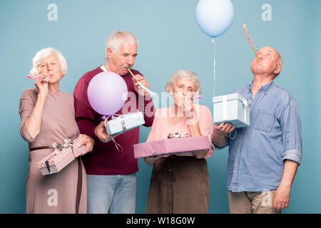 Two senior couples with giftboxes blowing whistles while having birthday fun Stock Photo