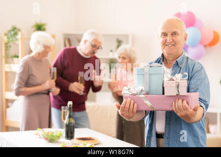 Senior smiling man with several packed gifts looking at you at home party with friends Stock Photo