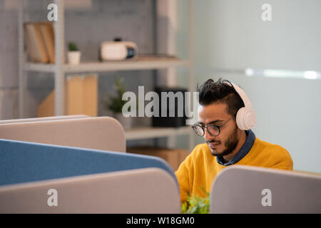 Serious thoughtful young Arabian office worker in eyeglasses sitting at table with partitions and listening to audio file in headphones while working Stock Photo